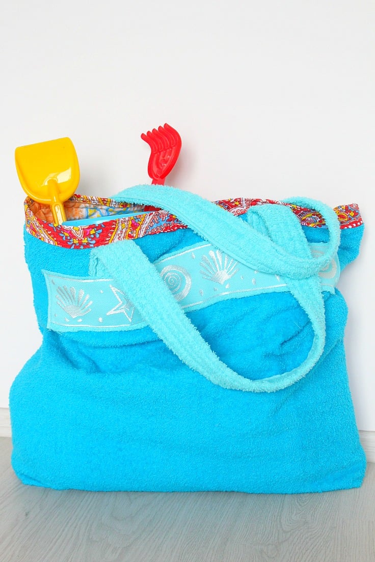 A towel bag for beach in two shades of blue, filled with beach toys for kids, sitting against a white wall. 