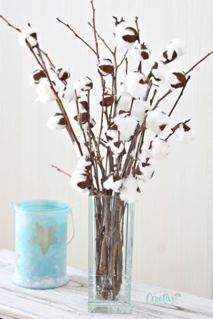 DIY cotton stems with faux cotton branches, in a transparent vase next to a blue candle holder.