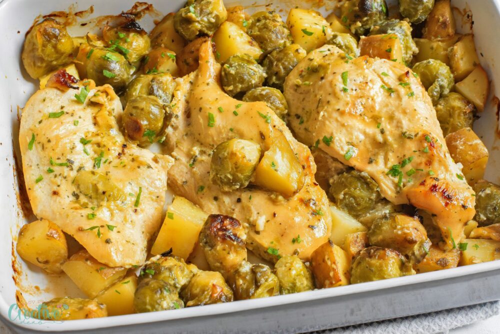 Juicy honey Dijon chicken breast surrounded by roasted Brussels sprouts and potatoes in a cozy baking dish.